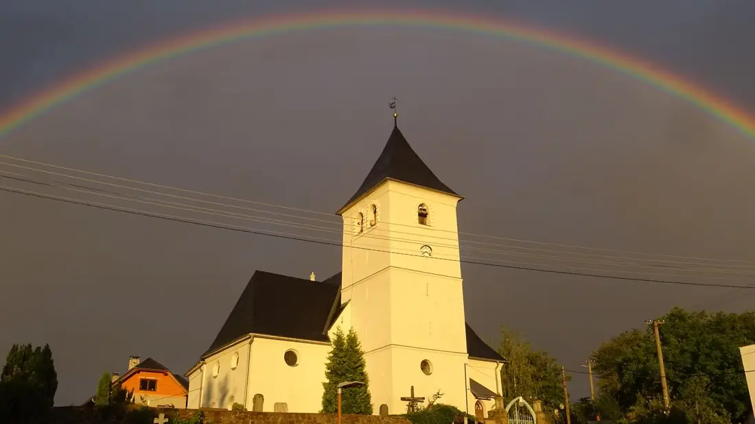 Kostelní věž obklopená střechami domů s duhou na obloze po bouřce.