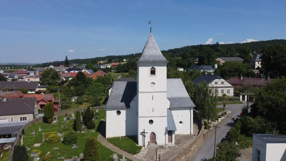 Bílá kostelní věž jako dominanta nad střechami obce při pohledu z výšky.
