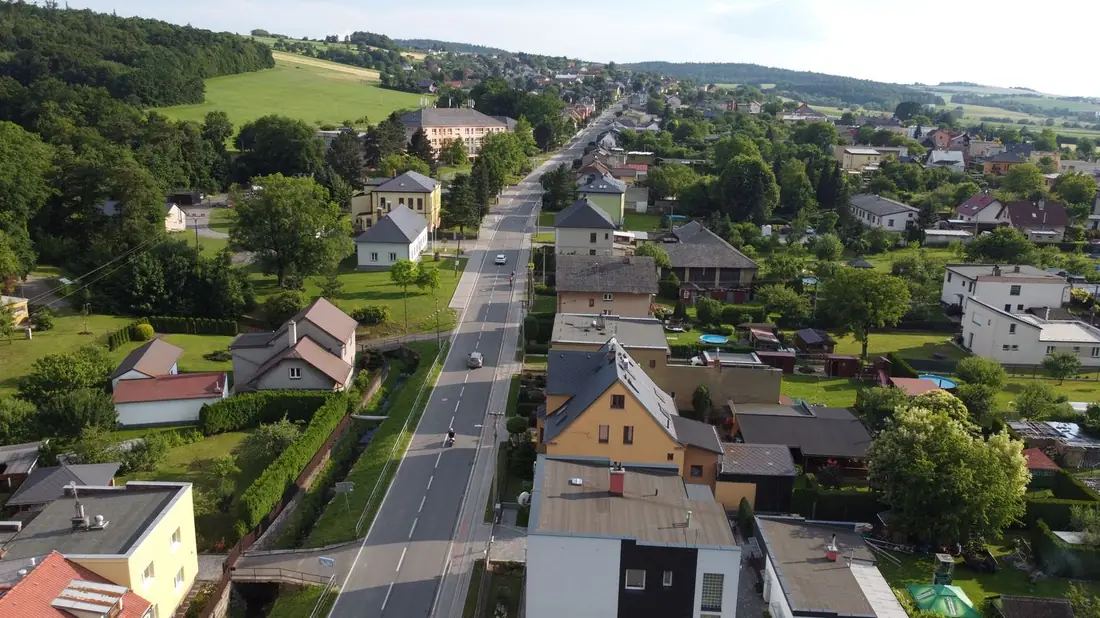 Průjezdní silnice obcí lemovaná řadou rodinných domů a zahrad.